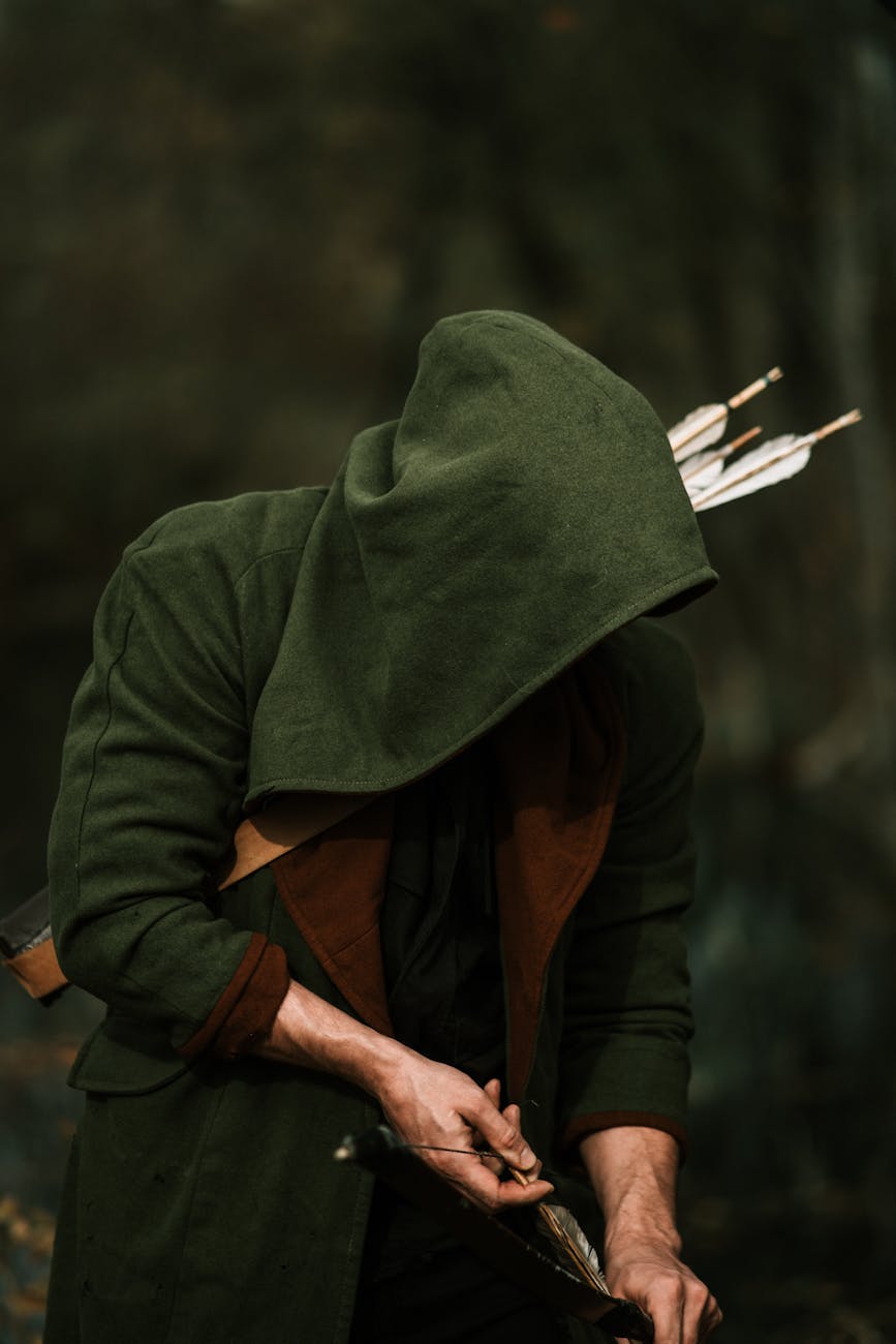man wearing a hood holding a bow and arrows in a forest